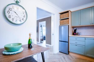 Eine moderne Küche mit einem Holztisch, einer grünen Schüssel und einer Champagnerflasche. Eine große Wanduhr ist sichtbar.