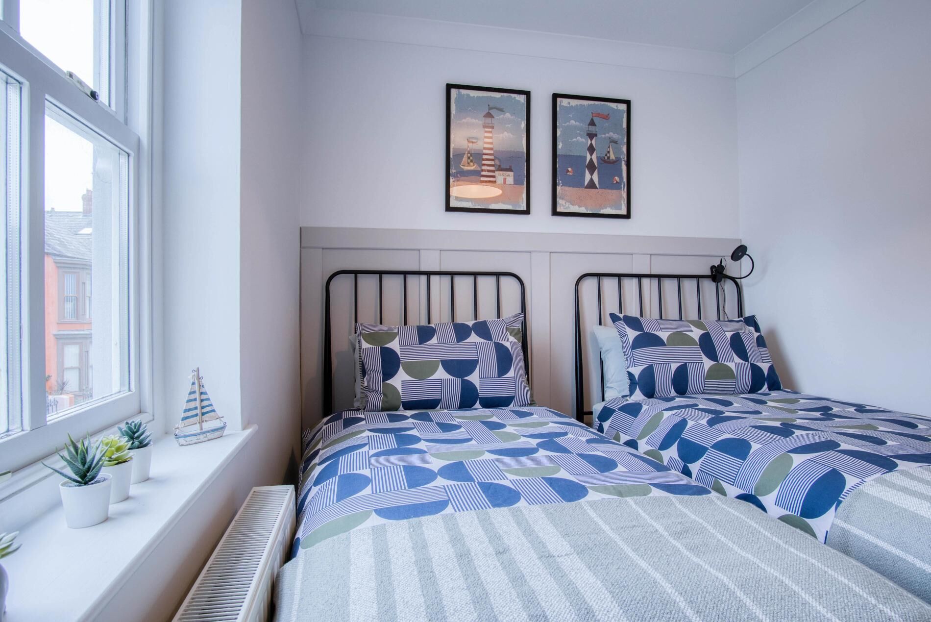 A bedroom with two beds, a radiator, a window with plants, and two framed pictures on the wall.