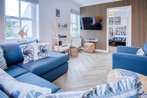 Un salon moderne avec un canapé bleu, un téléviseur à écran plat fixé sur un mur recouvert de panneaux en bois, et un lit avec une couvre-lit à motifs géométriques en arrière-plan.