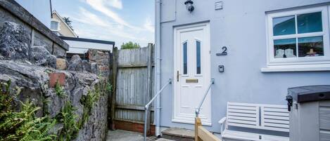 A narrow stone wall with a wooden gate, a white door with a number plate, and a small wooden bench outside a building with a light blue exterior.