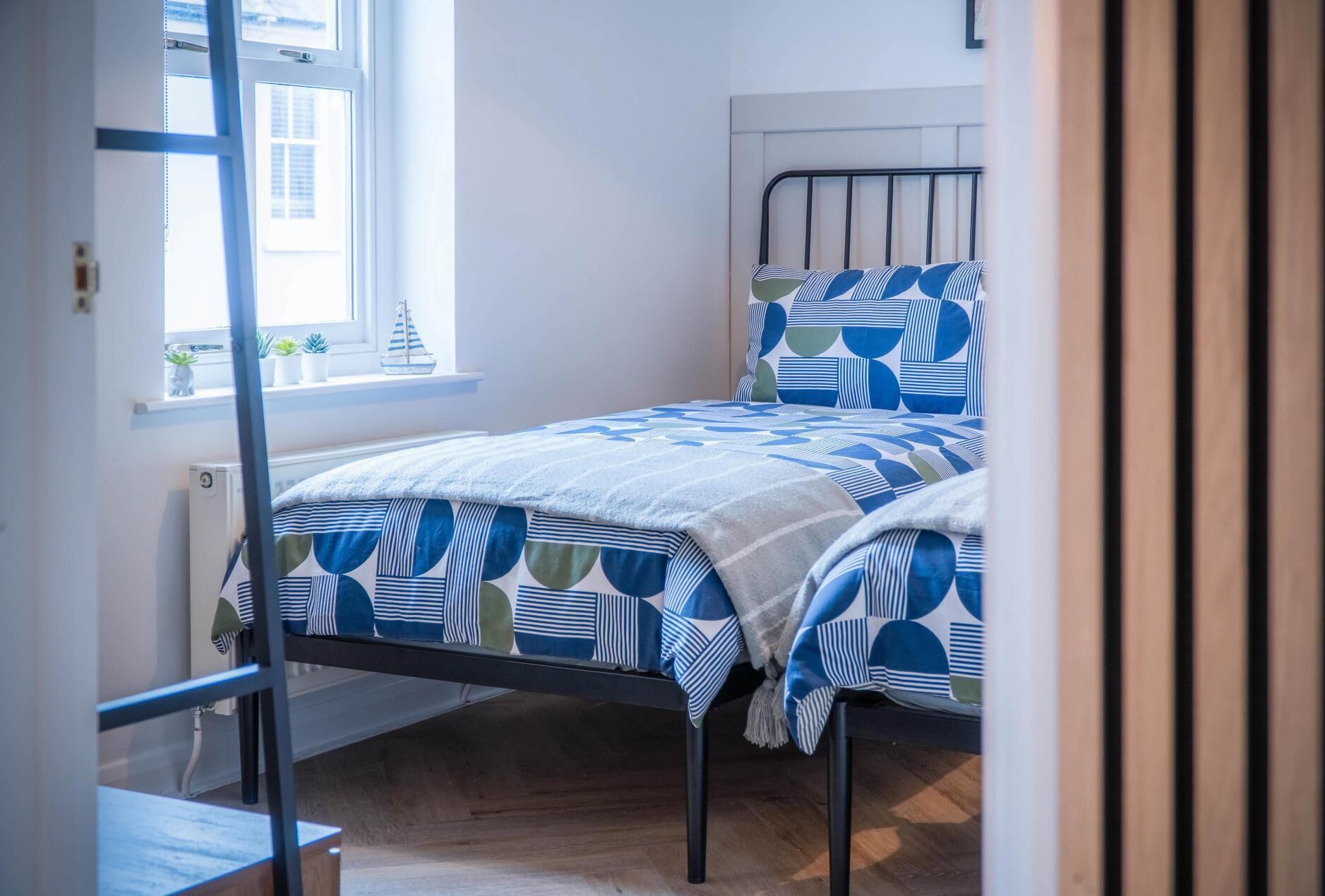 Un lit simple avec une couvre-lit à motifs géométriques, une fenêtre donnant sur des immeubles et un radiateur fixé au mur.