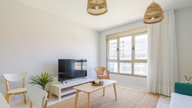 A modern living room with a flat-screen TV, wicker pendant lights, a white sofa, a dining table, and a large window with curtains. - CITY FAMILY AUDITORIUM (Las Palmas de Gran Canaria)