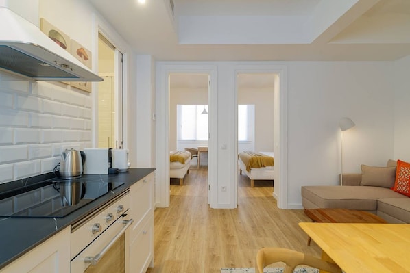 Une cuisine moderne avec des meubles blancs et un comptoir noir, un coin repas avec une table et des chaises en bois, et un salon avec un canapé et une lampe.