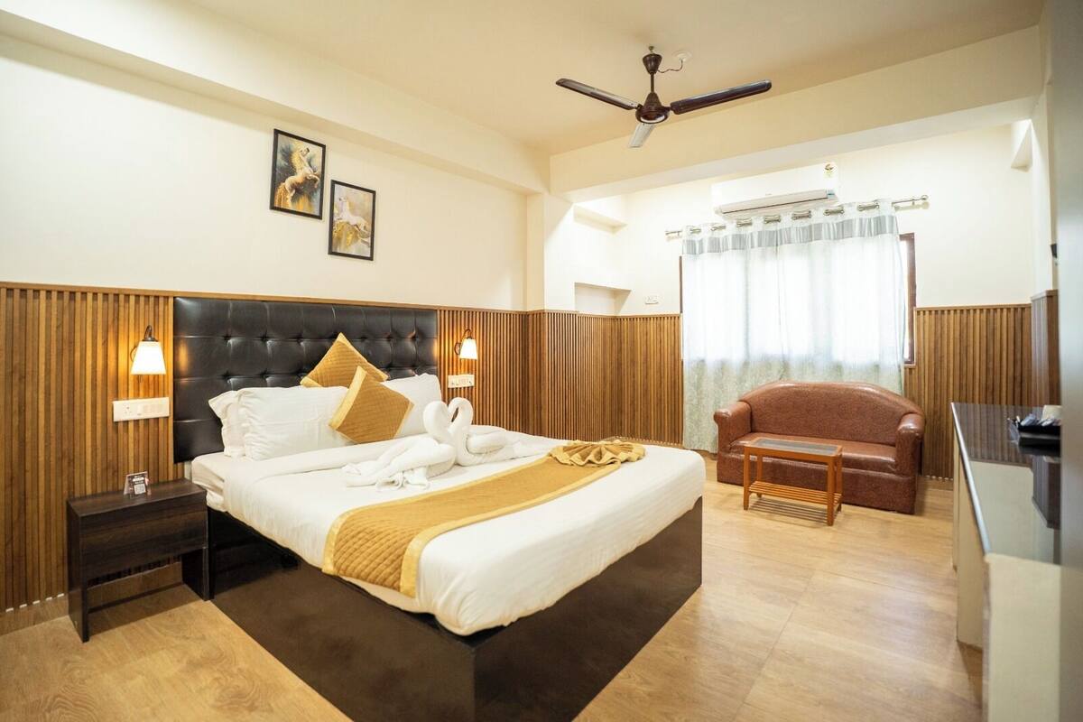 Quarto de hotel com uma cama grande, cabeceira de madeira, duas luminárias fixadas na parede, ventoinha no teto, sofá de couro marrom e piso de madeira.
