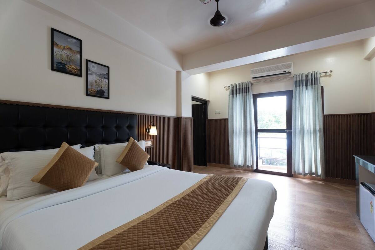 Quarto de hotel com uma cama grande, cabeceira, mesa de cabeceira, luminária e janela com cortinas.