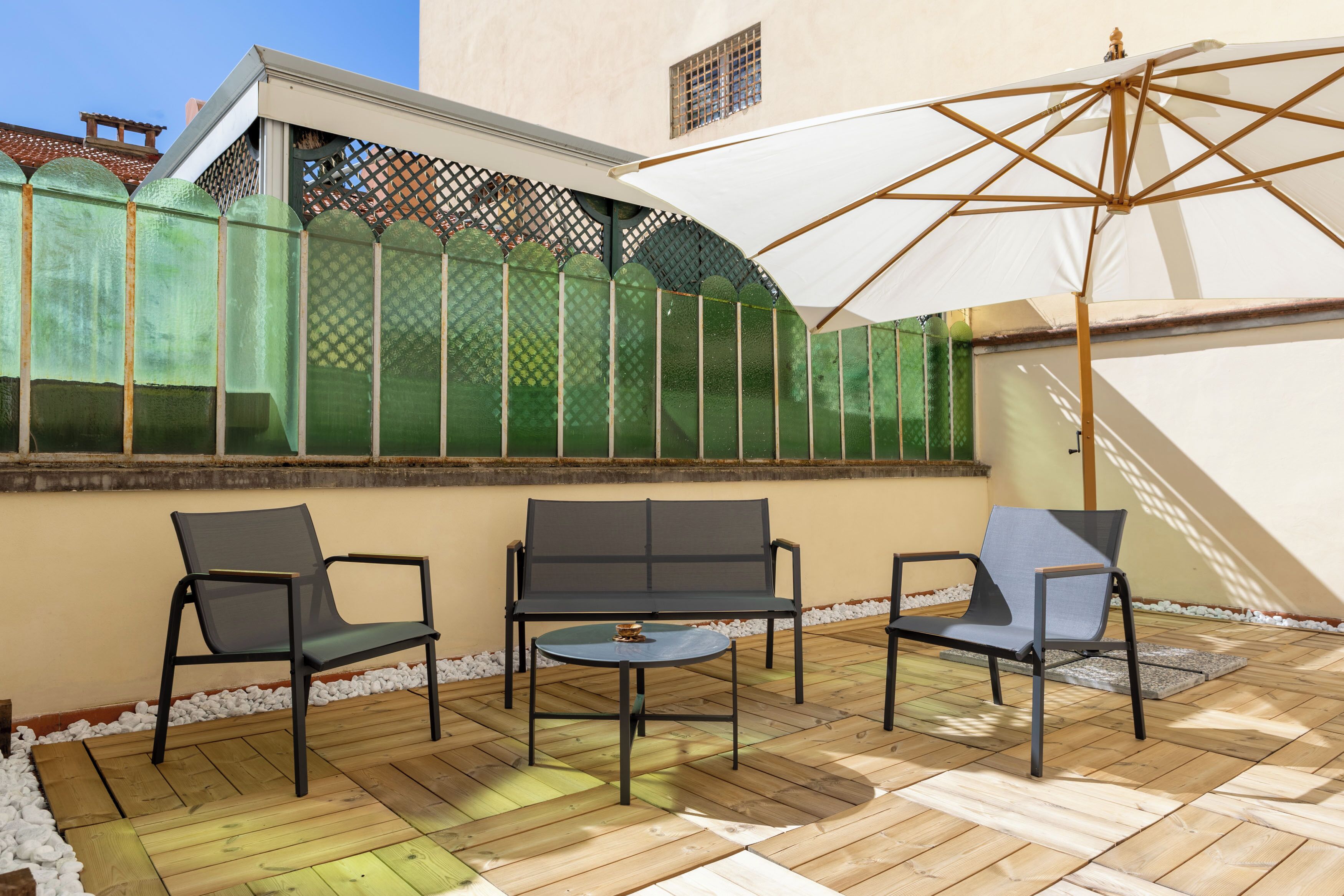 Um pátio externo com um deck de madeira, dois sofás cinzentos, uma mesa redonda pequena e um guarda-sol branco.