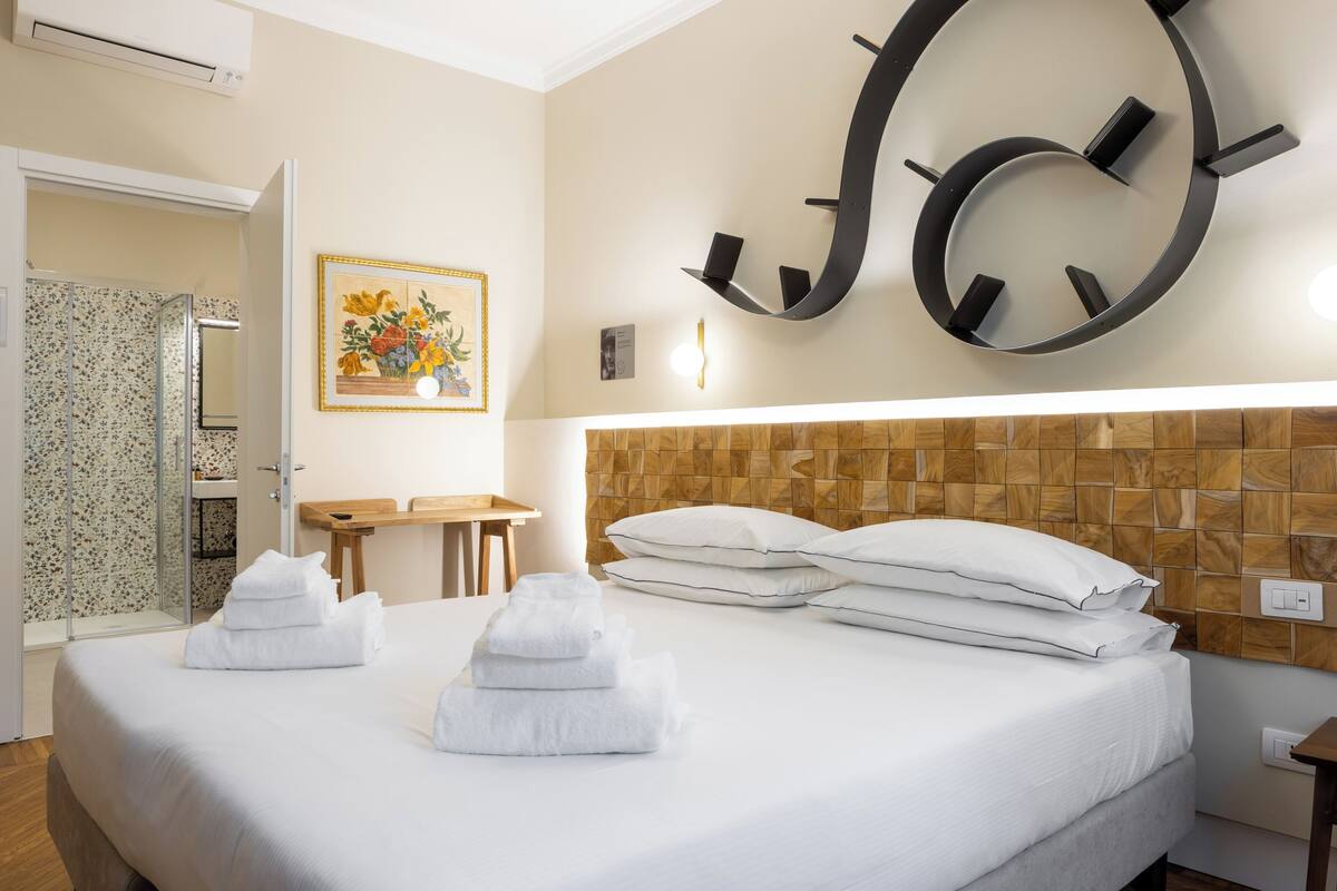 A hotel room with a neatly made bed, a wooden headboard, a wall-mounted clock, and a small table with a chair.
