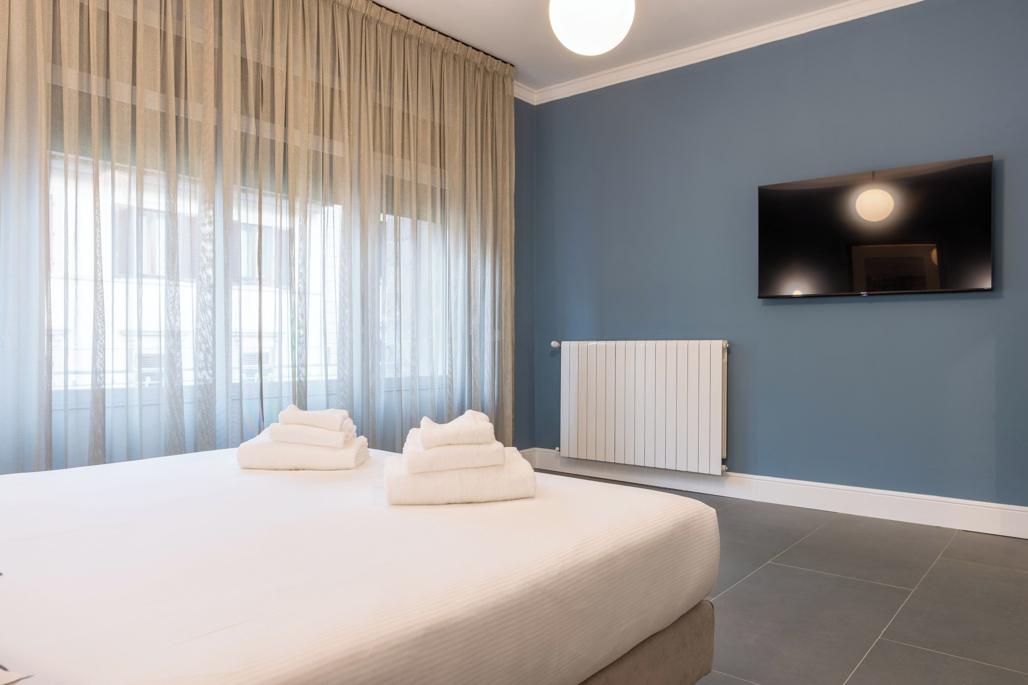 A modern hotel room with a large bed, a flat-screen TV mounted on the wall, and a large window with sheer curtains.