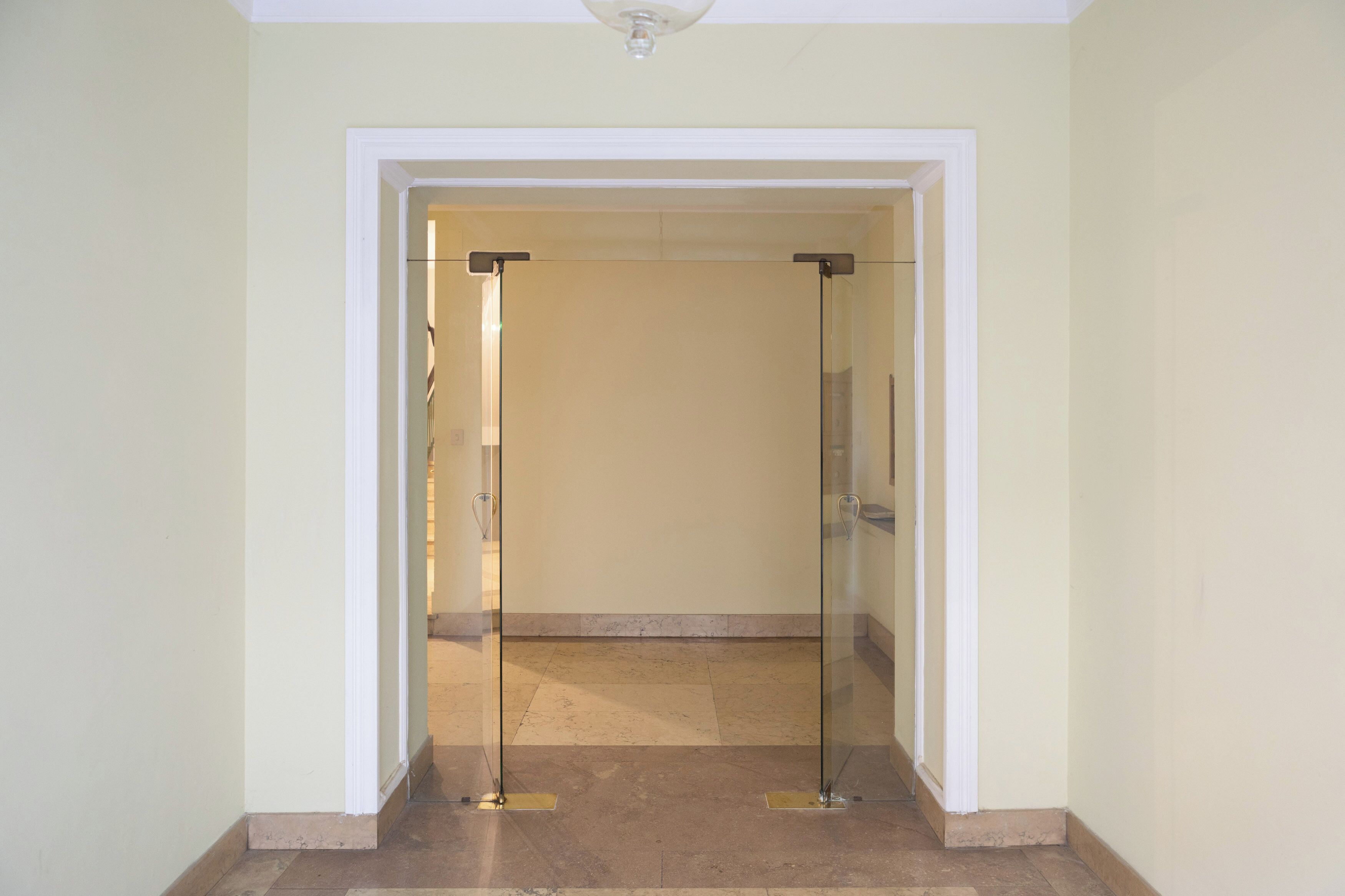 A hallway with a glass door leading to another room. The walls are painted in a light color, and the floor is tiled.