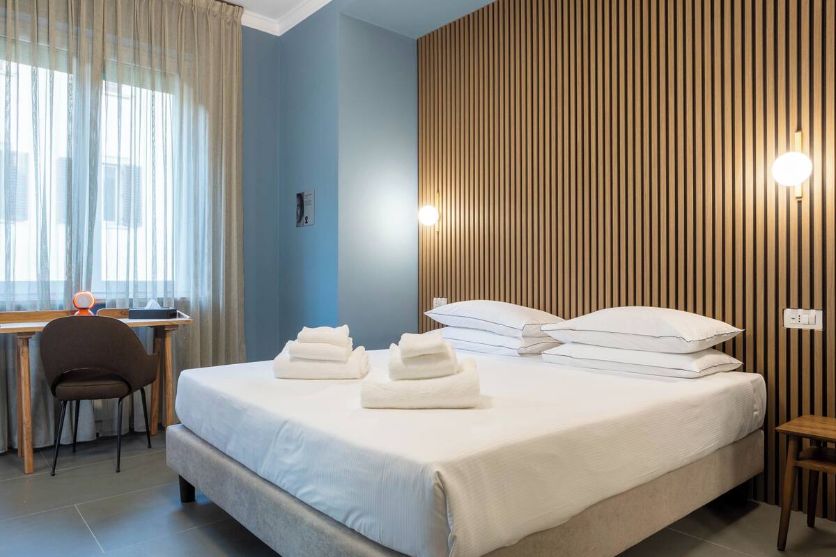 A neatly made bed with white linens and pillows, a wooden desk with a chair, and a wall with vertical wooden slats and two wall-mounted lights.