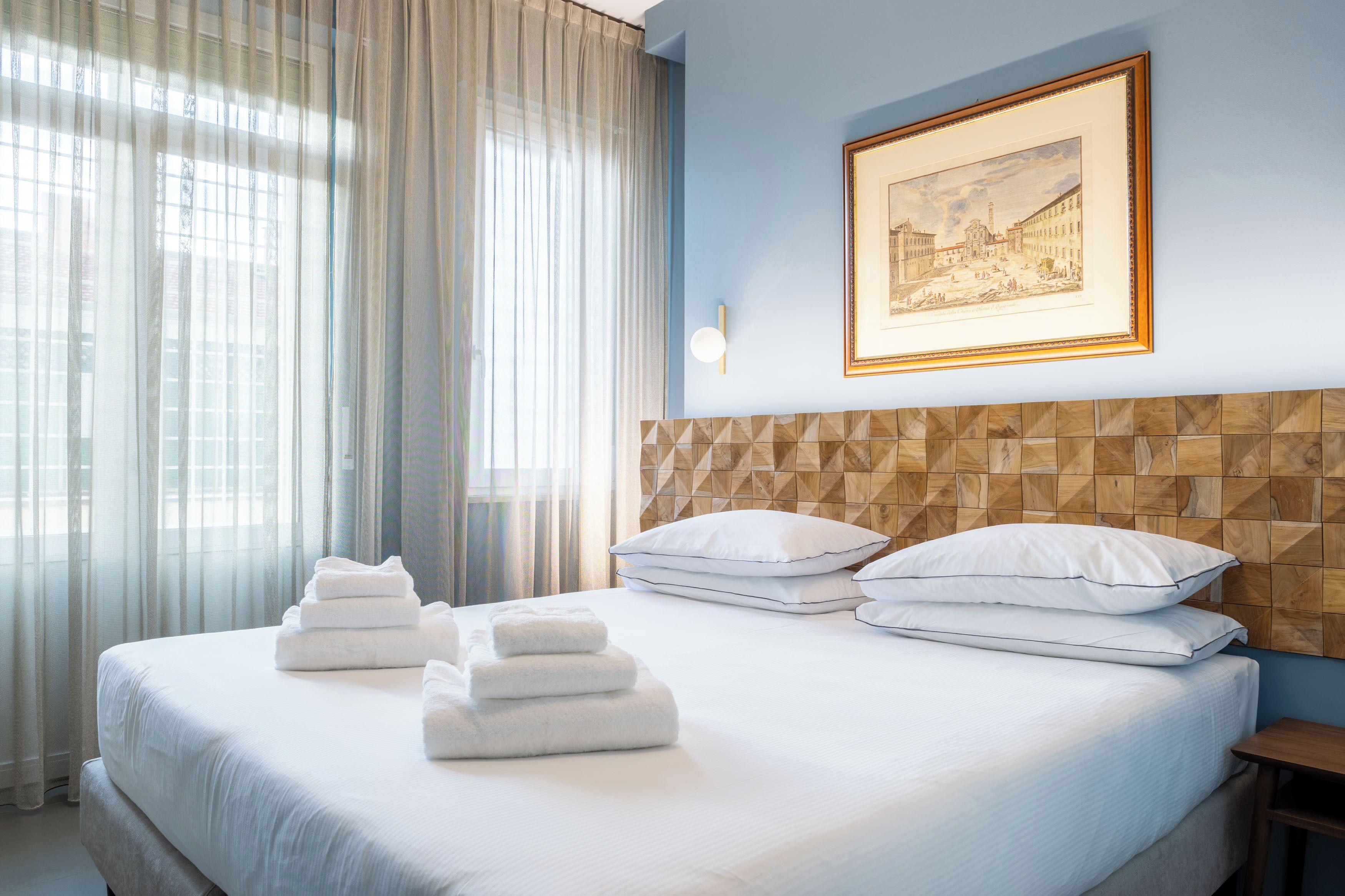 A hotel room with a large bed, white linens, a geometric-patterned headboard, a framed artwork on the wall, and sheer curtains.