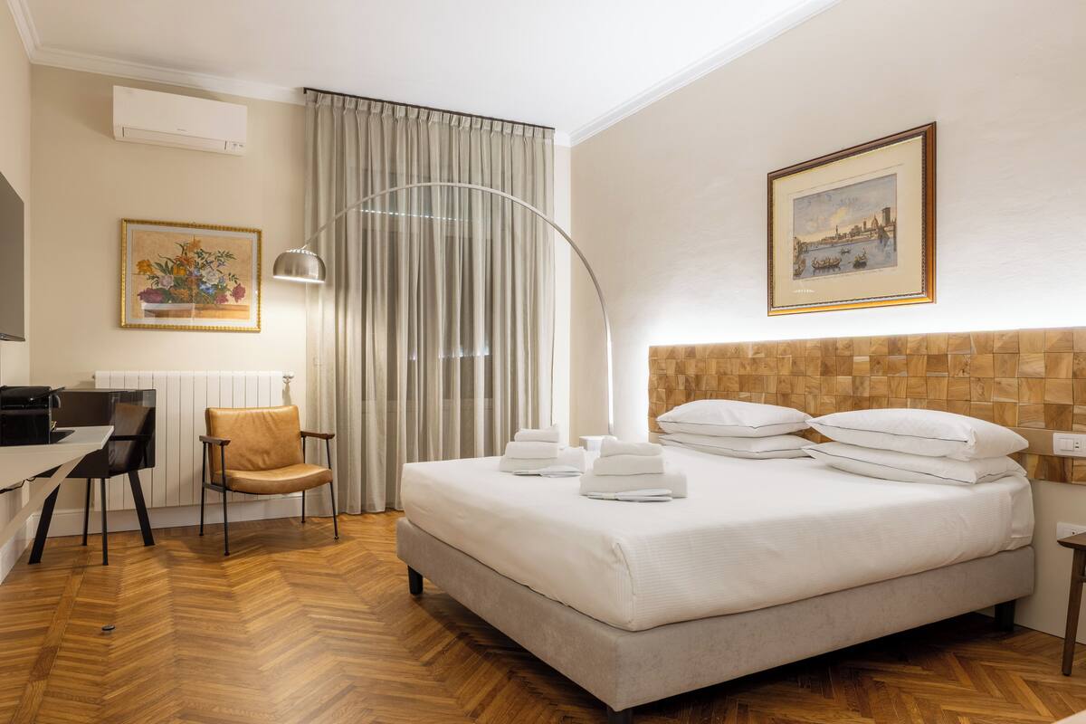 A hotel room with a bed, a desk, a chair, a lamp, and a painting on the wall.