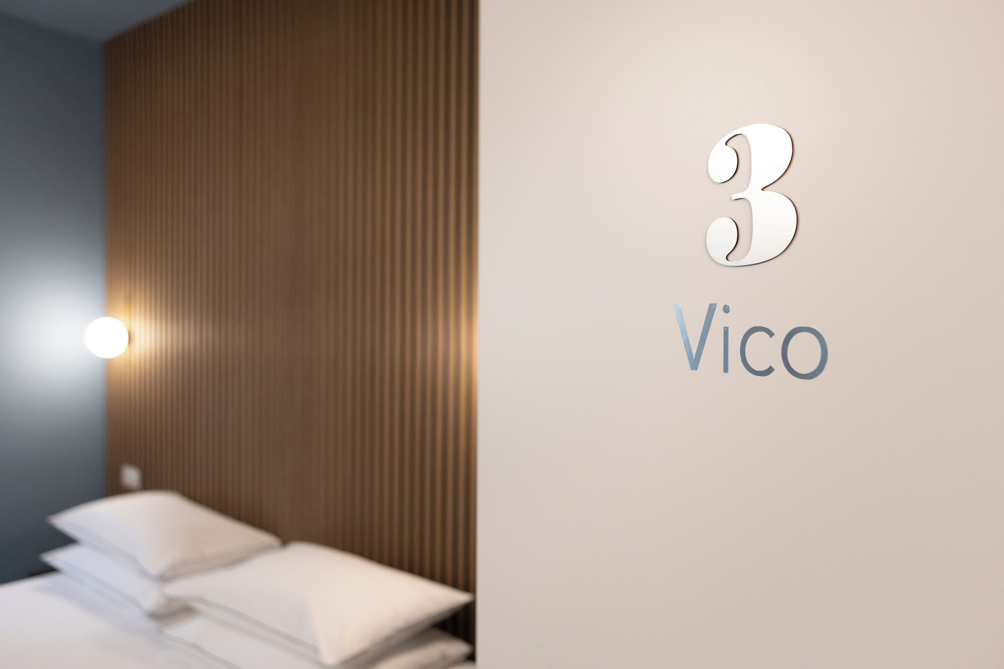 A hotel room with a bed, a wall-mounted light, and a number '3' with the name 'Vico' on the wall.