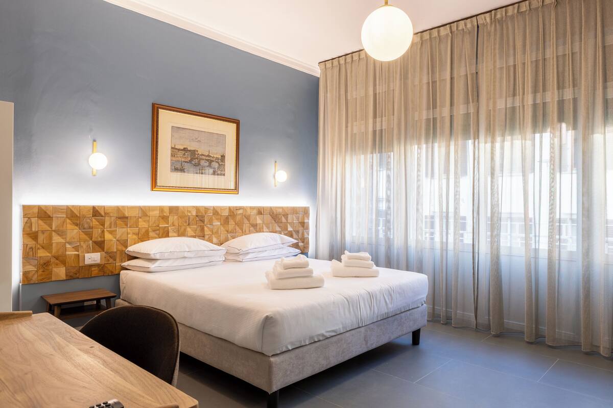 A hotel room with a bed, a wooden headboard, a framed picture on the wall, and sheer curtains.