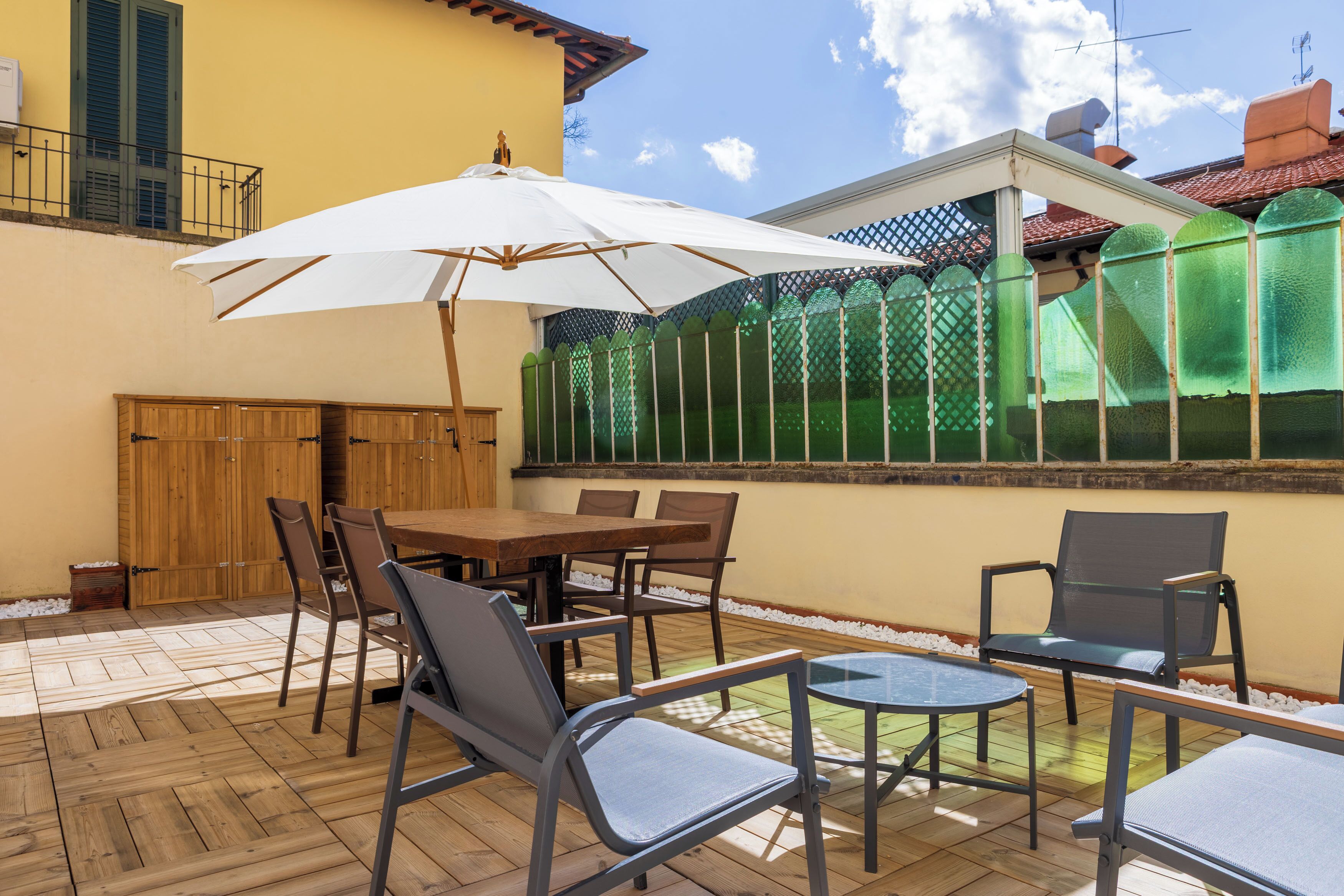 An outdoor patio with a wooden table and chairs, a white umbrella, and a green fence.