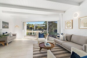 Un vaste salon doté d’une grande porte-fenêtre coulissante qui donne sur un balcon offrant une vue sur un plan d’eau et des bâtiments. Le salon est aménagé avec un canapé, une table basse et un fauteuil.
