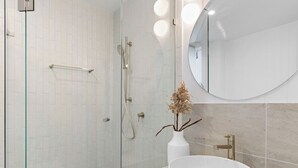 Une salle de bain moderne avec une douche à l’italienne en verre, un lavabo blanc avec un robinet doré et un miroir rond placé au-dessus du lavabo.