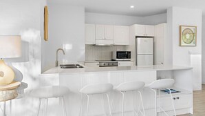 Une cuisine moderne avec des meubles blancs, un évier en acier inoxydable, un four à micro-ondes et un réfrigérateur. On y trouve des tabourets de bar blancs et une petite table avec une lampe.