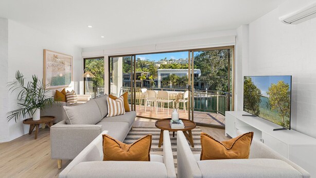 Un salon moderne avec un canapé, une table basse et un téléviseur. Il y a un balcon avec vue sur la mer et les bâtiments.