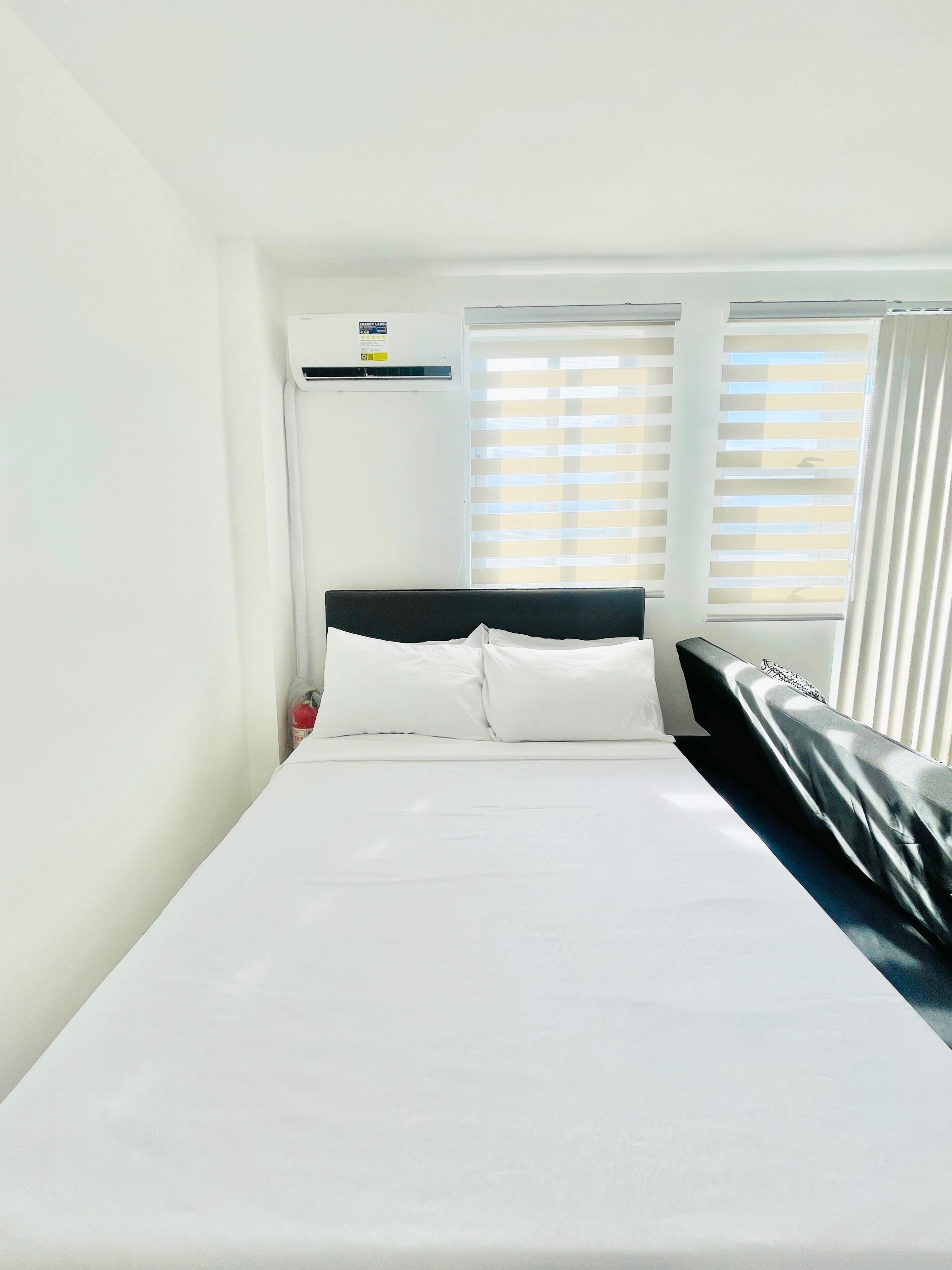 A hotel room with a bed, a headboard, a window with blinds, and a wall-mounted air conditioning unit.