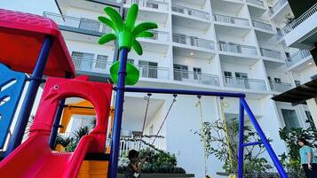 Un parque infantil con un tobogán rojo y columpios, rodeado de césped artificial, frente a un edificio de varios pisos con balcones.