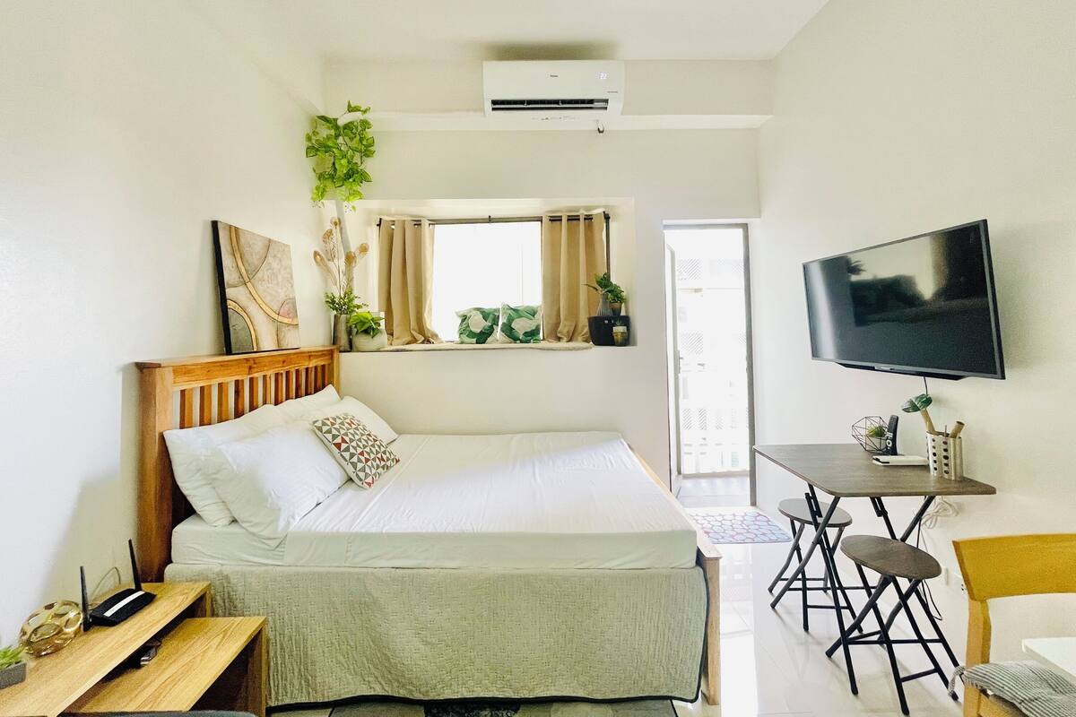 A bedroom with a bed, a desk, a chair, a TV, an air conditioner, and a window with curtains.