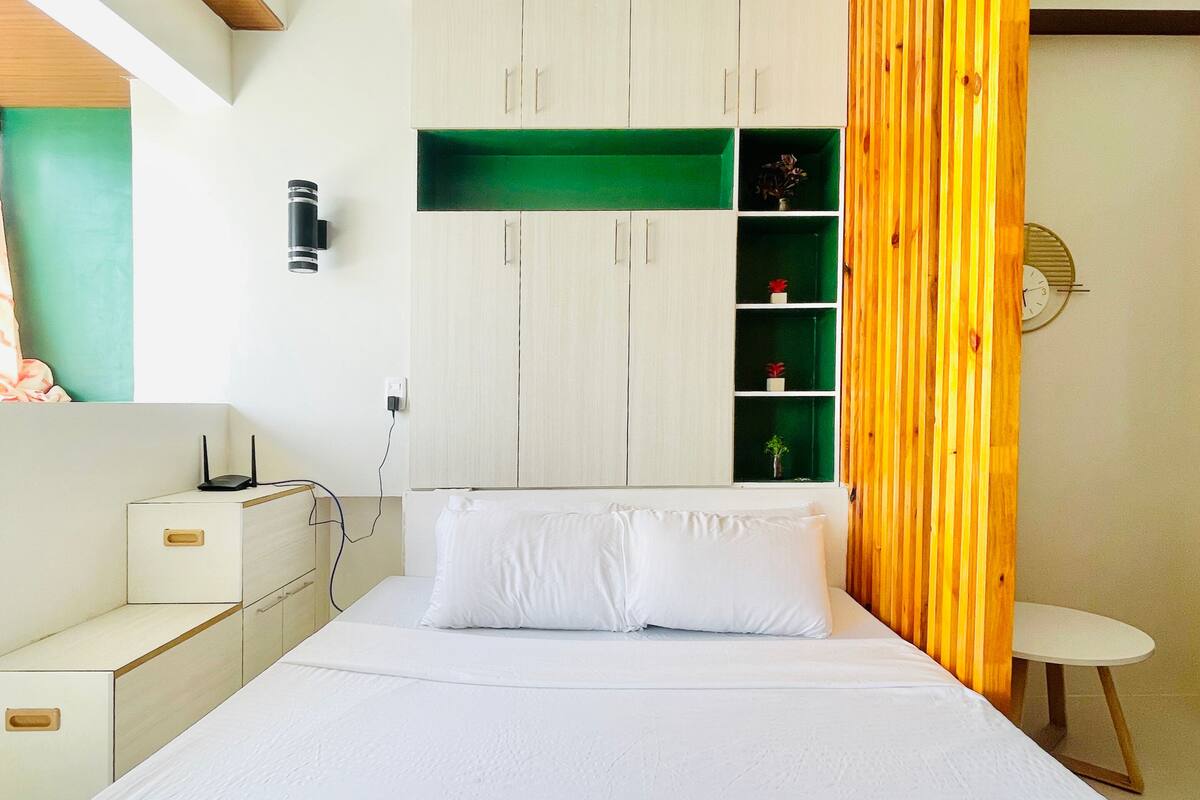 A modern bedroom with a bed, a nightstand, a dresser, a green shelf unit, and a wooden partition.