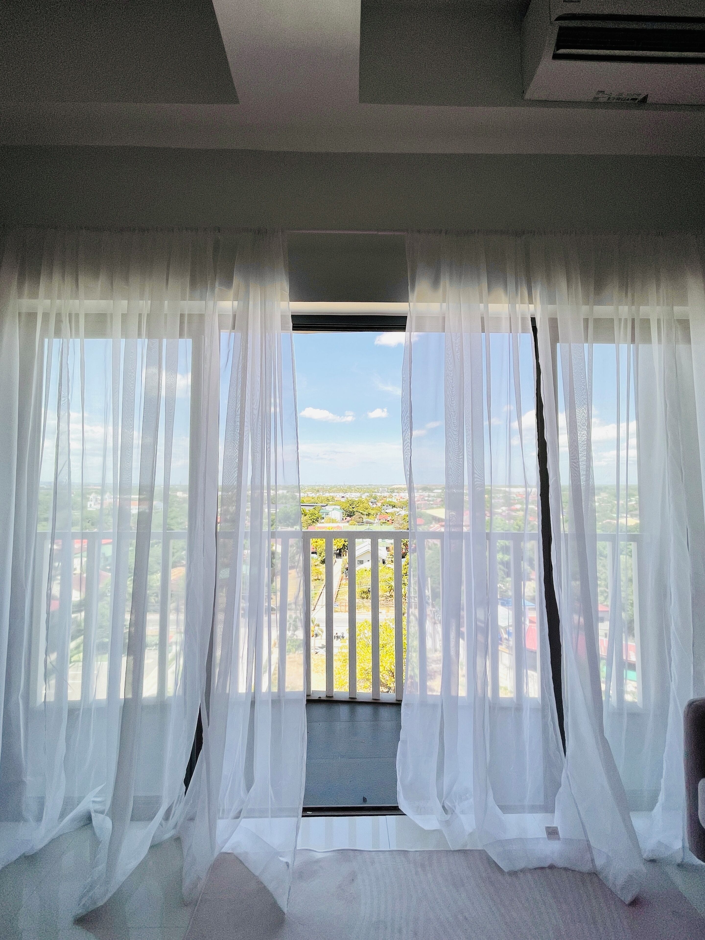 Een kamer met lichte witte gordijnen, een balkon met uitzicht op groen en gebouwen, en een raam met uitzicht op de lucht.