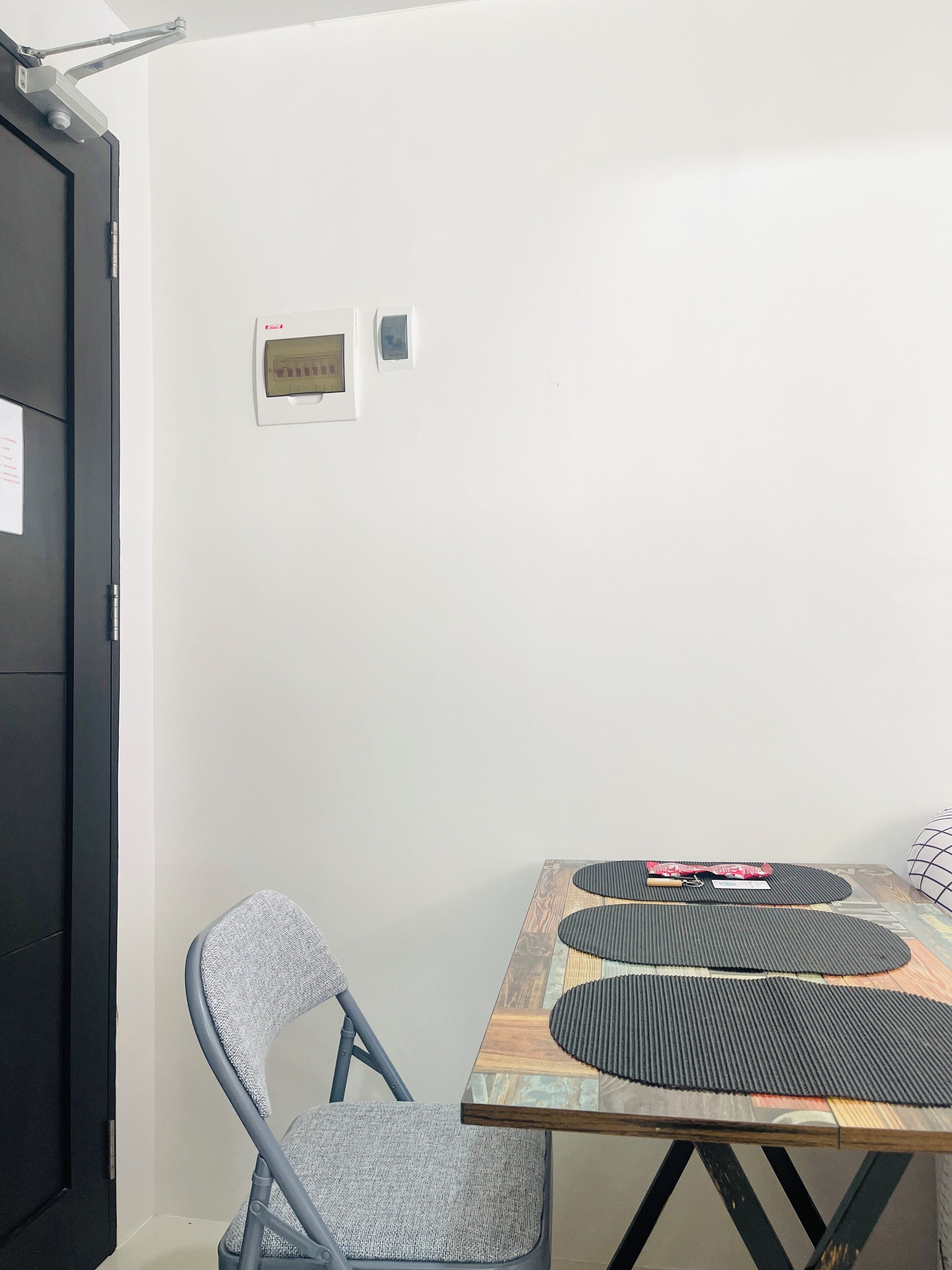A room with a wooden table, a grey upholstered chair, and a black door. There are two wall-mounted devices, one with a display and one with a control panel.
