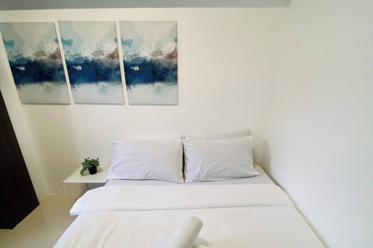 A neatly made bed with white linens and a rolled towel, a small bedside table with a plant, and three abstract paintings on the wall.