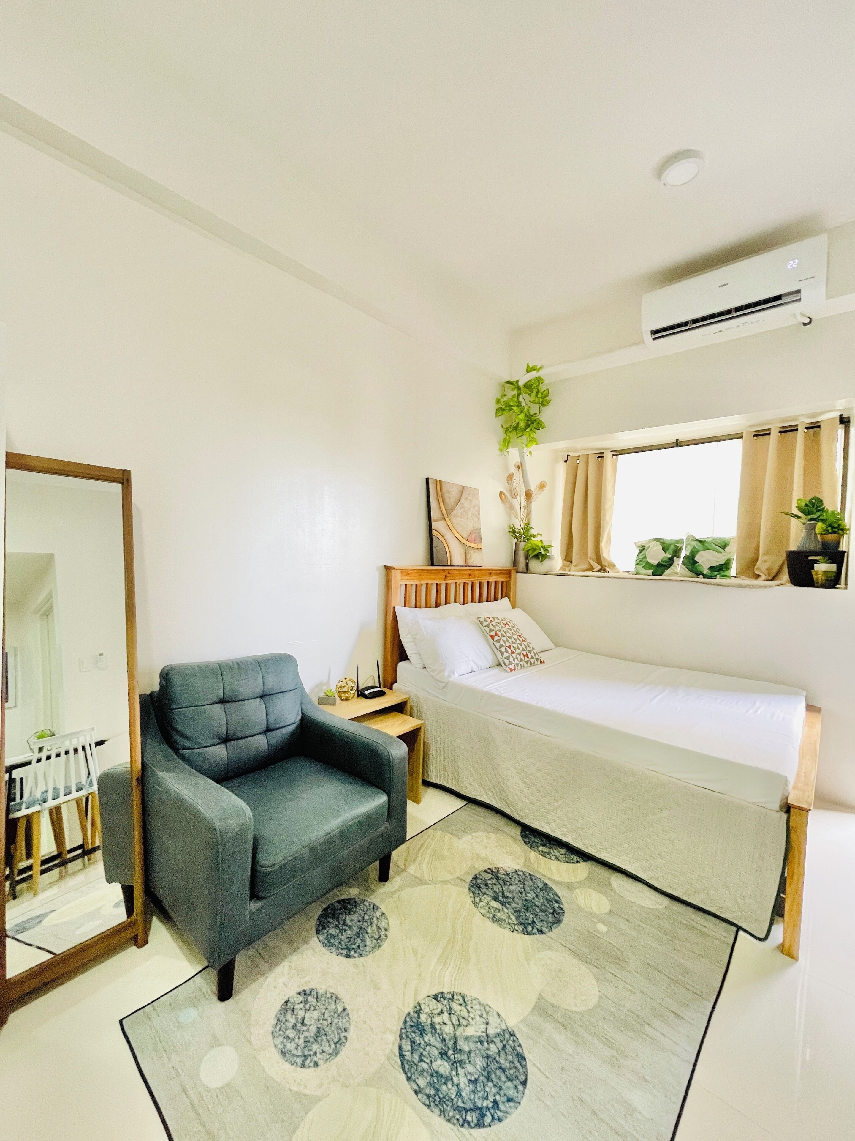 Un dormitorio con una cama individual, un sillón gris, una mesita de noche, un espejo y una ventana con cortinas.