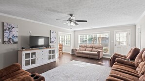 A spacious living room with a large flat-screen TV, a ceiling fan, and a beige sofa.