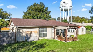 A house with a water tower in the background, a fenced yard, and a covered porch with wicker furniture.