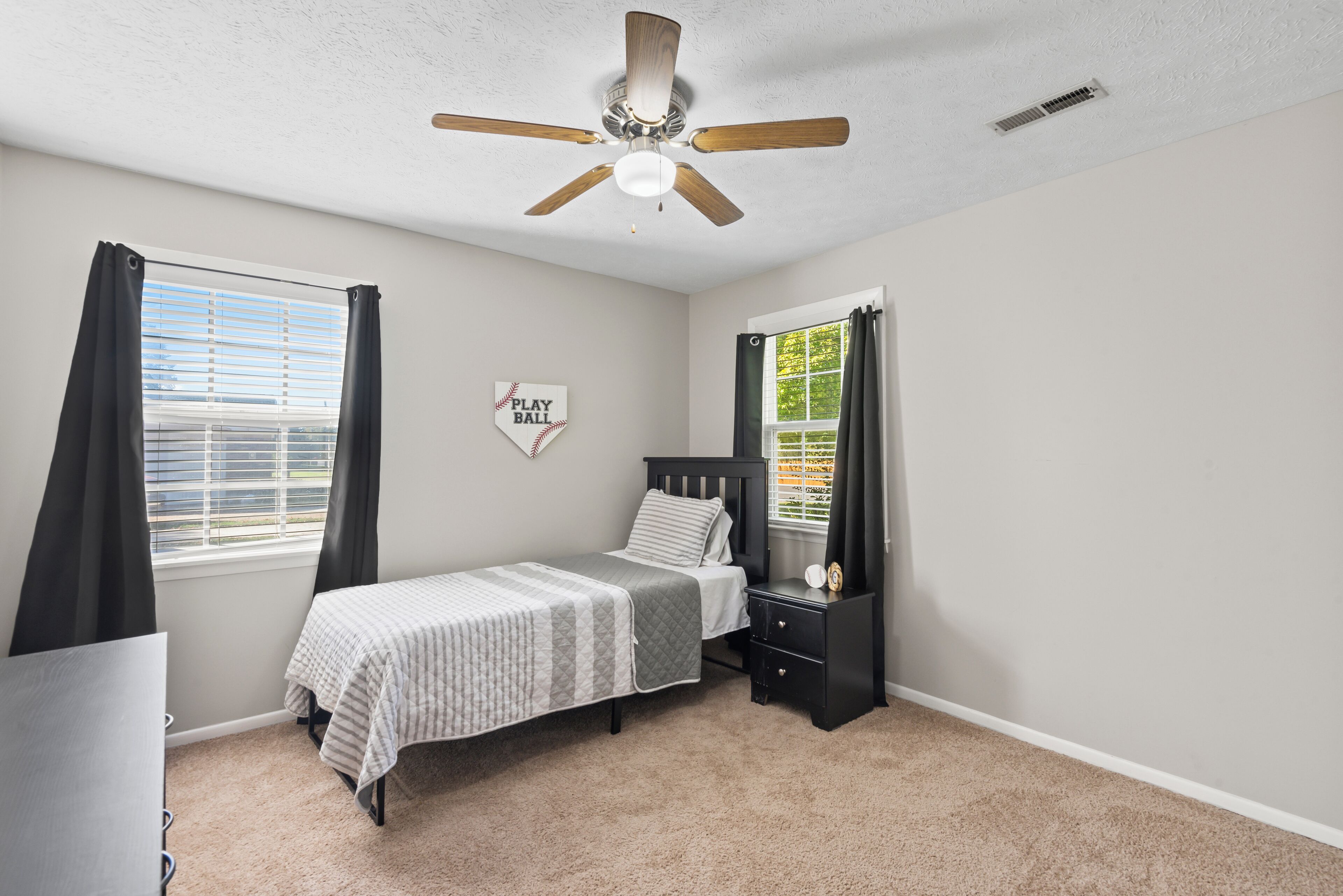 A bedroom with a bed, a nightstand, a ceiling fan, a window with curtains, and a wall-mounted decoration.