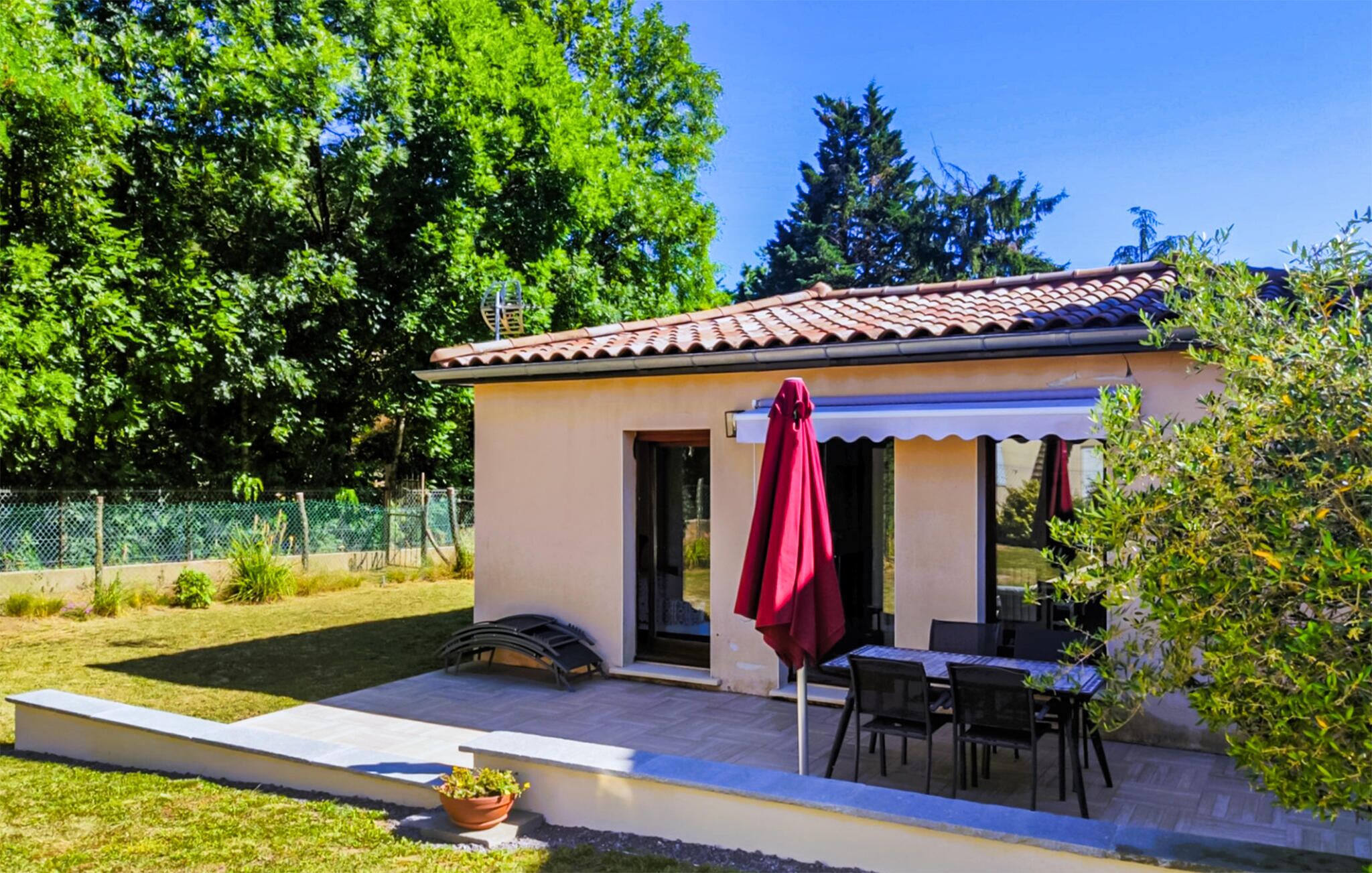 A small, single-story building with a tiled roof, a covered patio area with a table and chairs, and a potted plant.