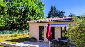 A small, single-story building with a tiled roof, a covered patio area with a table and chairs, and a potted plant.