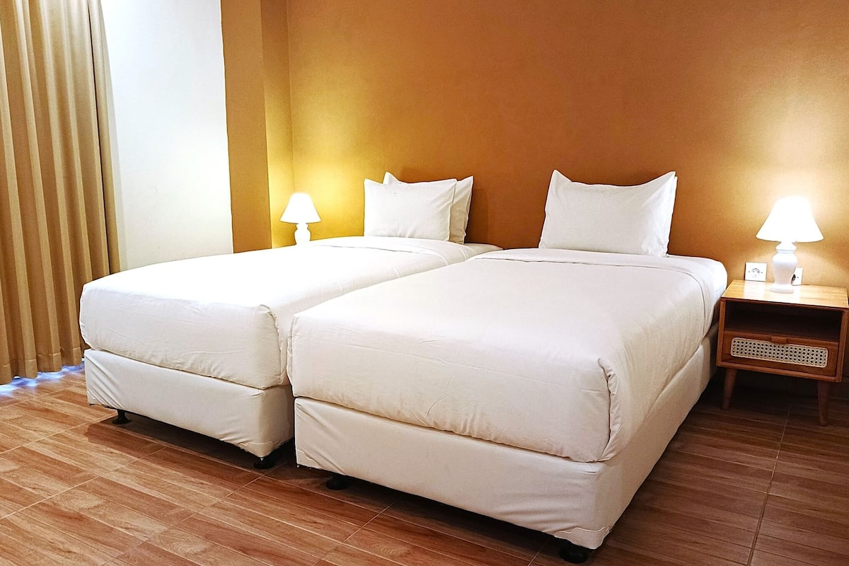 Une chambre d’hôtel avec deux lits, un plancher en bois, une table de chevet avec une lampe et une fenêtre avec des stores.