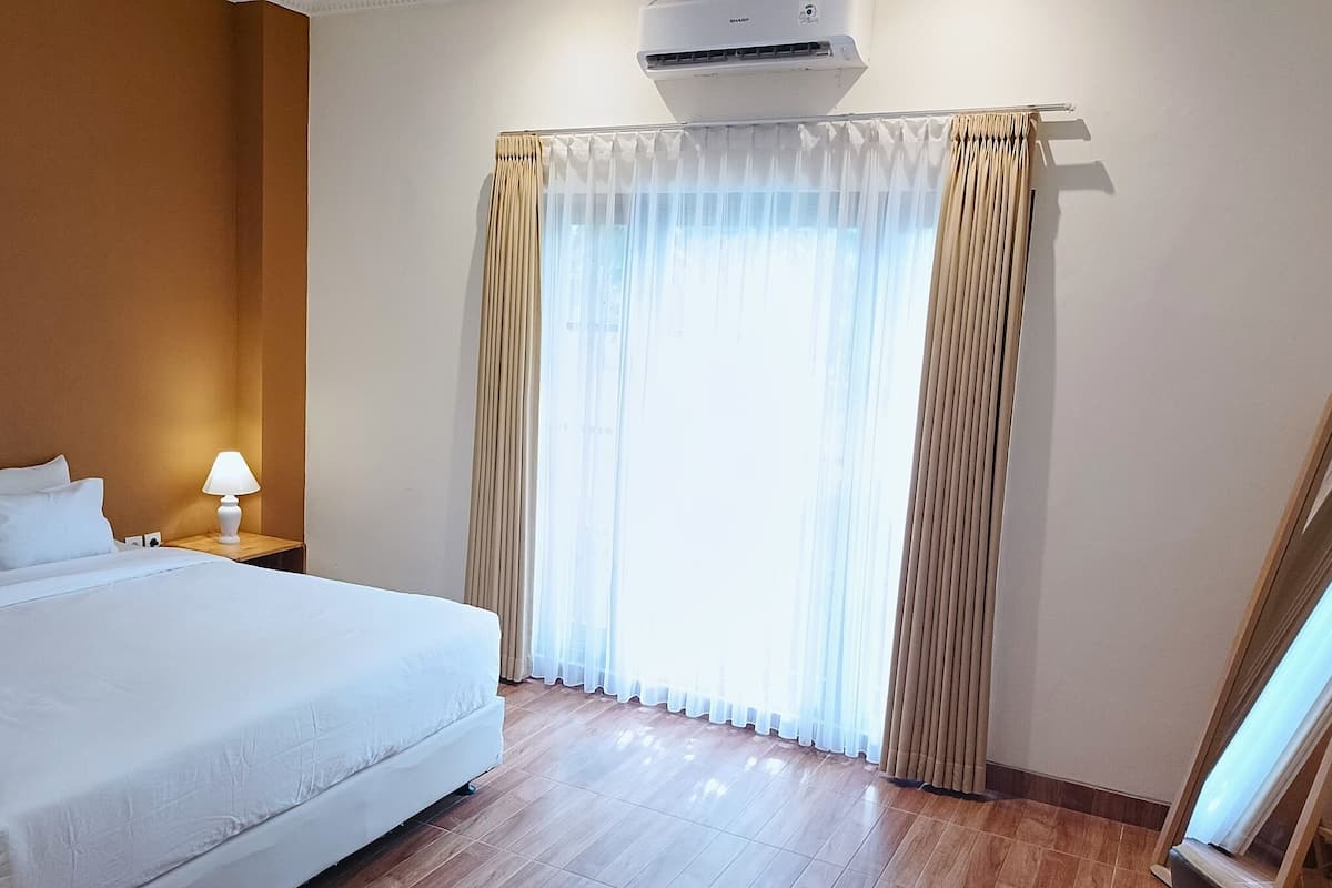 Une chambre d’hôtel avec un grand lit, une table de chevet avec une lampe, une fenêtre avec des rideaux légers et un climatiseur fixé au mur.