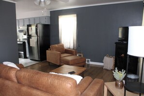 A living room with a sofa, a coffee table, and a television. There is a kitchen area with cabinets and a refrigerator.