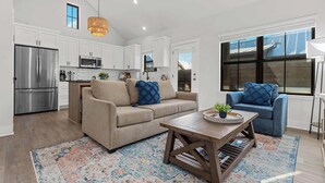 A modern kitchen with white cabinets, a stainless steel refrigerator, and a microwave. There is a beige sofa with blue cushions and a blue armchair. A wooden coffee table with a tray of items is in the center.