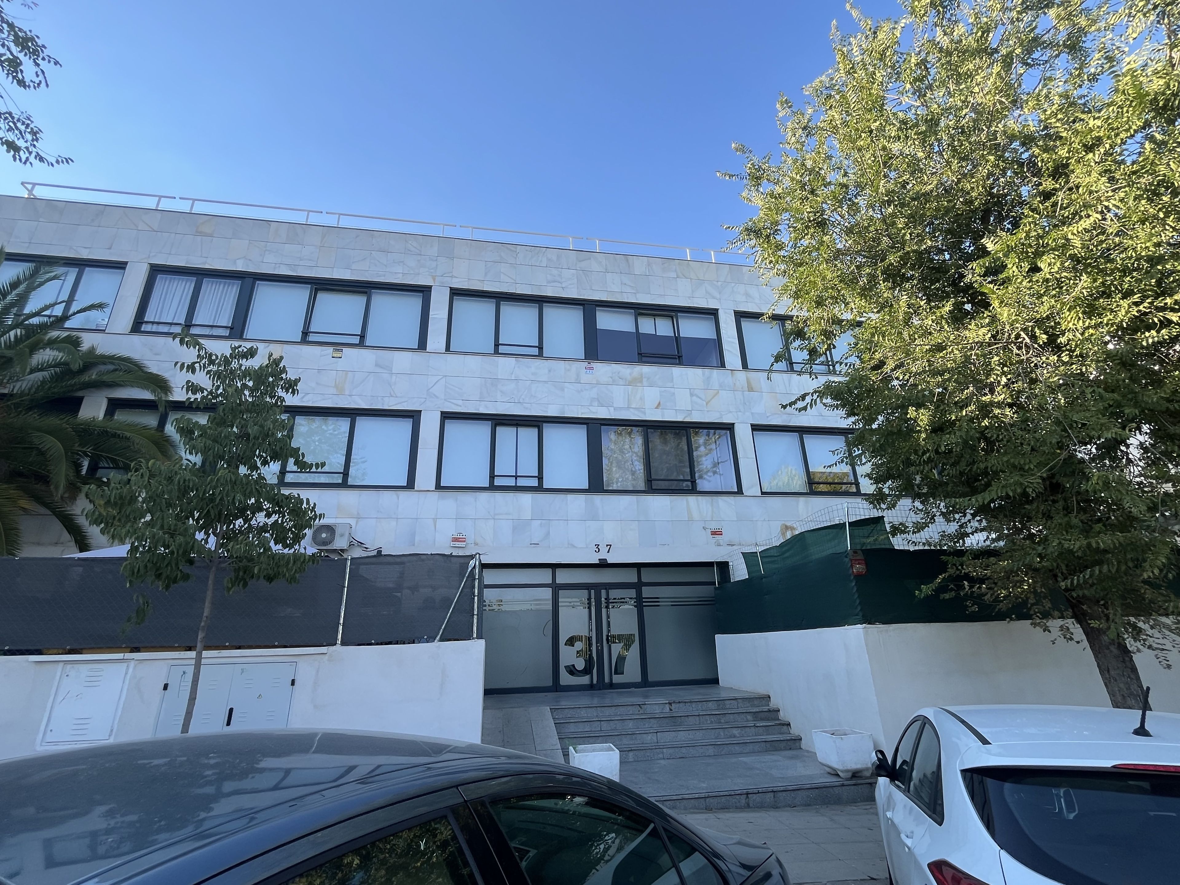 Un edificio a più piani con facciata moderna, ingresso delimitato da cancello e area parcheggio con auto.