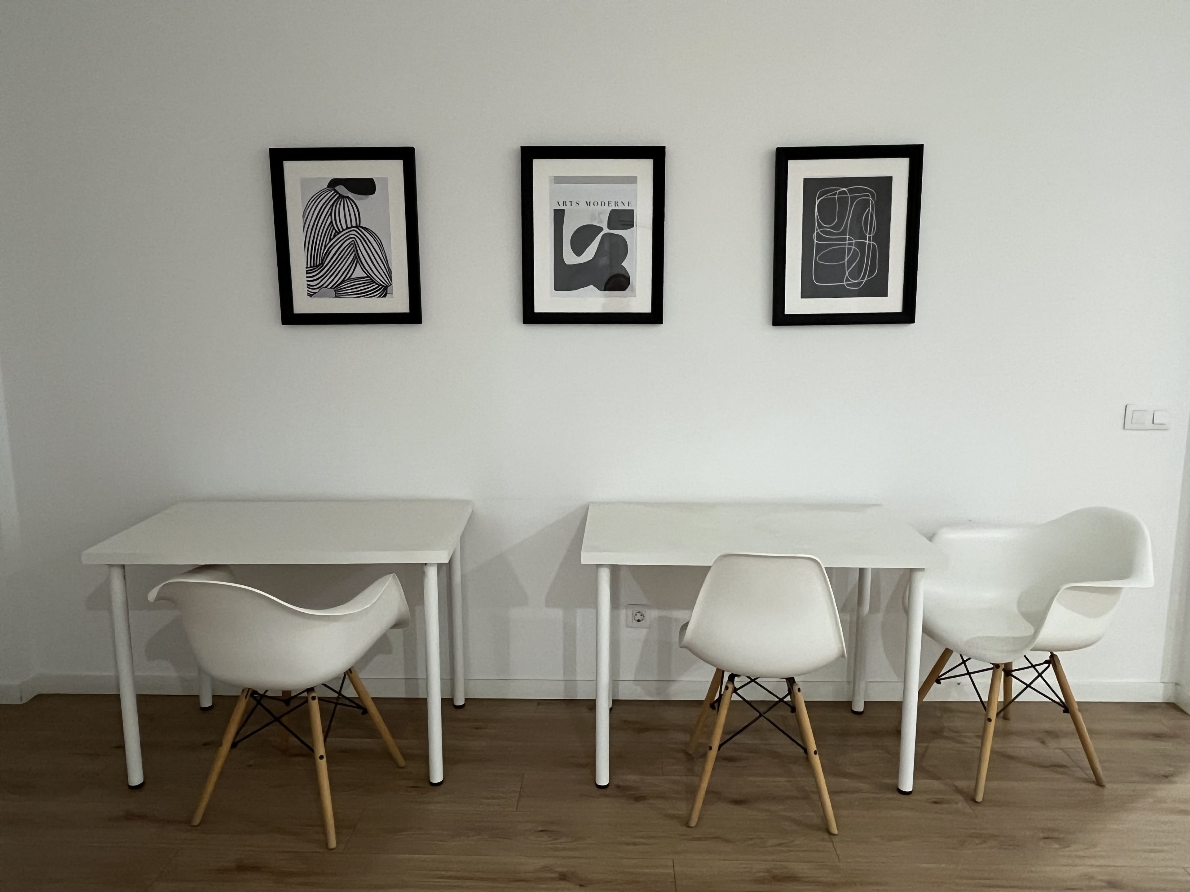 A room with a white table and two white chairs with wooden legs, a white floor, and three framed artworks on the wall.