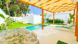 A covered outdoor area with a swimming pool, green lawn, and lounge chairs.