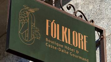 Letrero que dice 'FOLKLORE Boutique Hotel & Casse-Dalle Gourmand', con una ilustración de un pájaro y uvas.