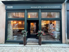 La entrada del Boutique Hotel & Casse-Dalle Gourmand Folklore, con un amplio ventanal que muestra un diseño de pájaro y frutas, dos plantas en macetas y una puerta de acceso visible.