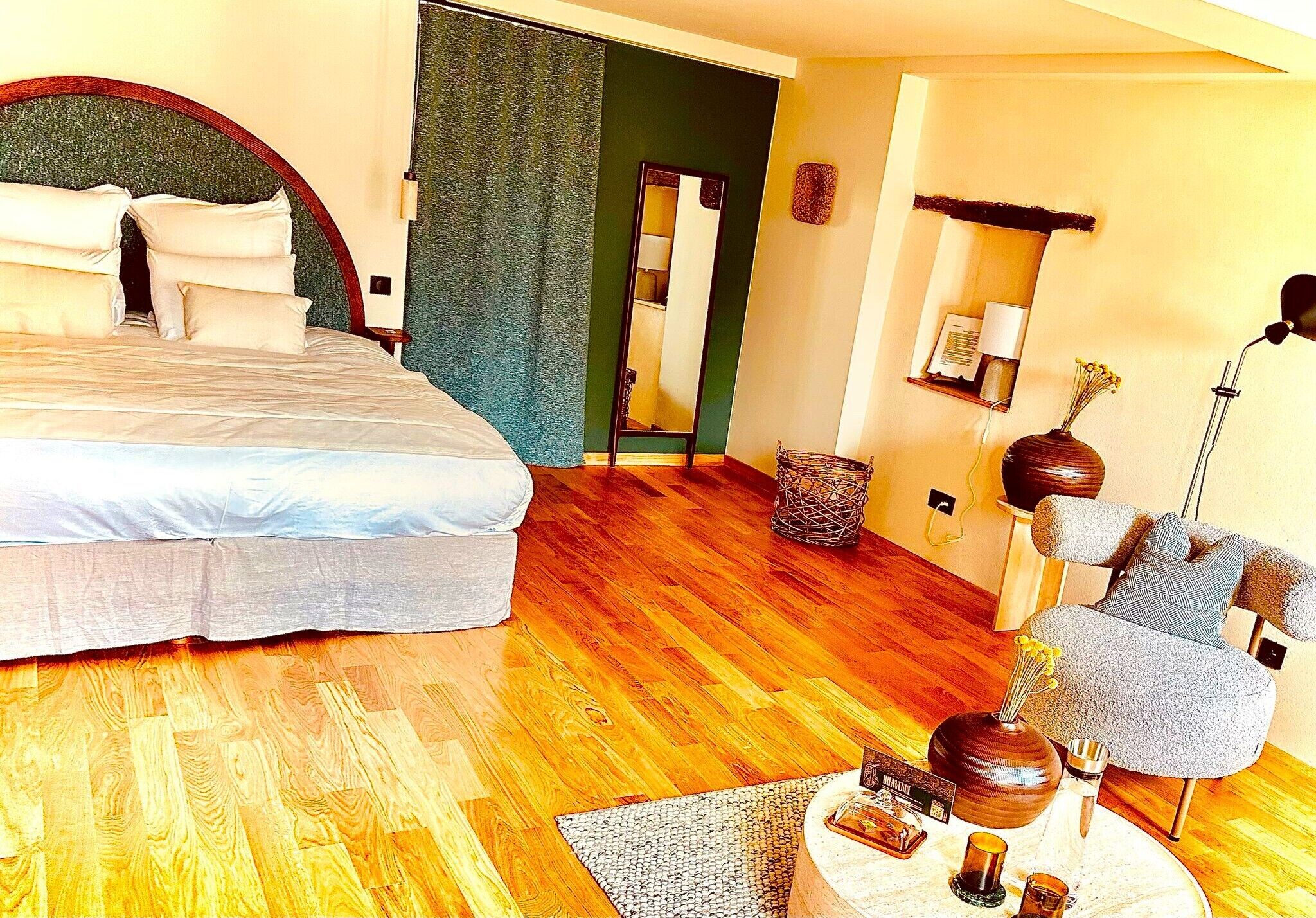 A hotel room with a large bed, a wooden floor, a sitting area with a chair and a small table, and a mirror on the wall.