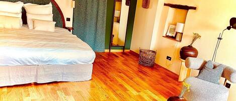 A hotel room with a large bed, a wooden floor, a sitting area with a chair and a small table, and a mirror on the wall.