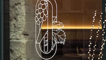 Ein Fenster mit einem Rebstock- und Vogelmotiv, eine Steinsäule und ein runder Tisch draußen.