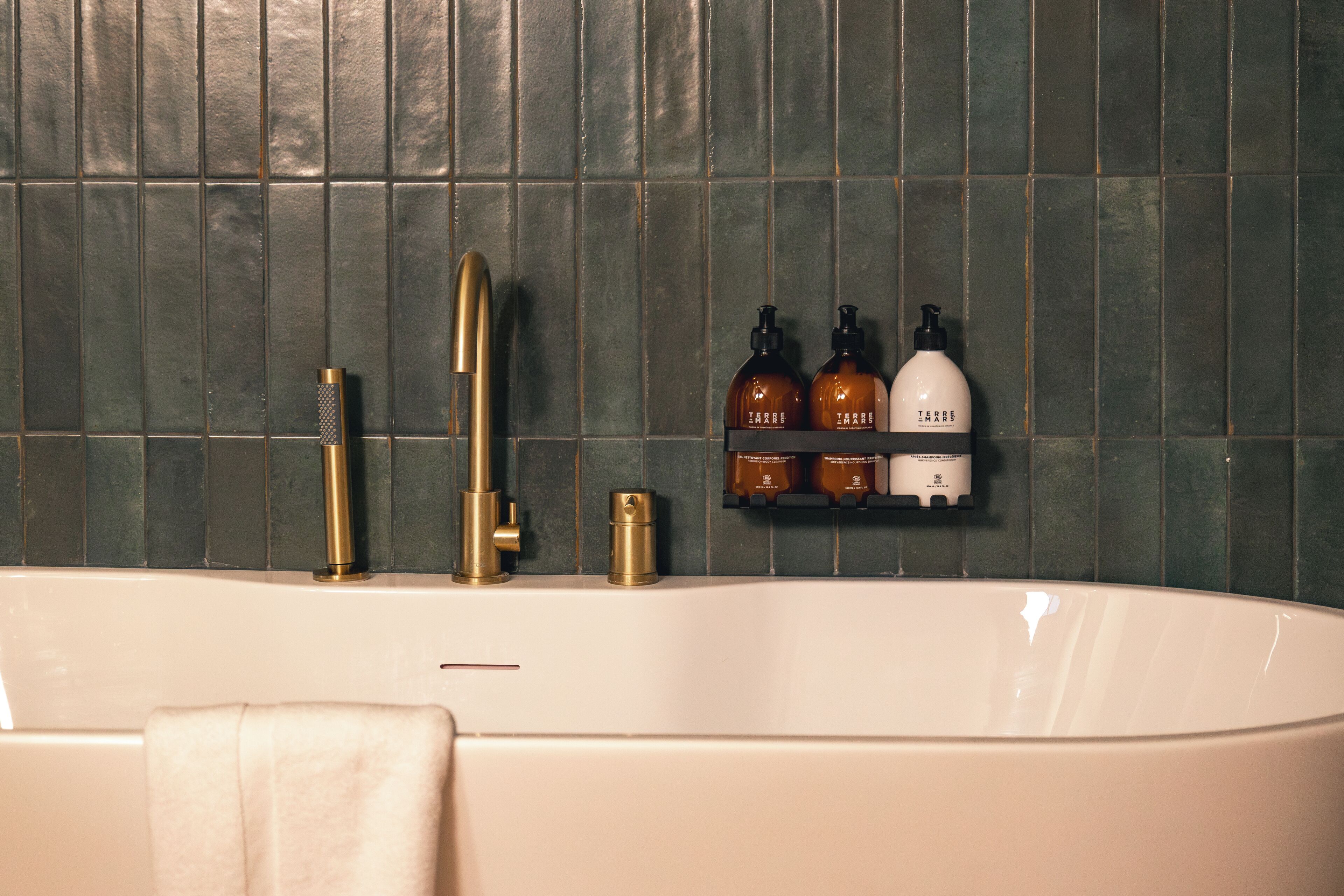 A bathroom with a freestanding bathtub, a gold faucet, and a towel hanging on the side. There are two brown bottles and a white bottle on a shelf above the tub.