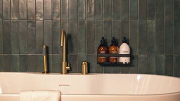 A bathroom with a freestanding bathtub, a gold faucet, and a towel hanging on the side. There are two brown bottles and a white bottle on a shelf above the tub.